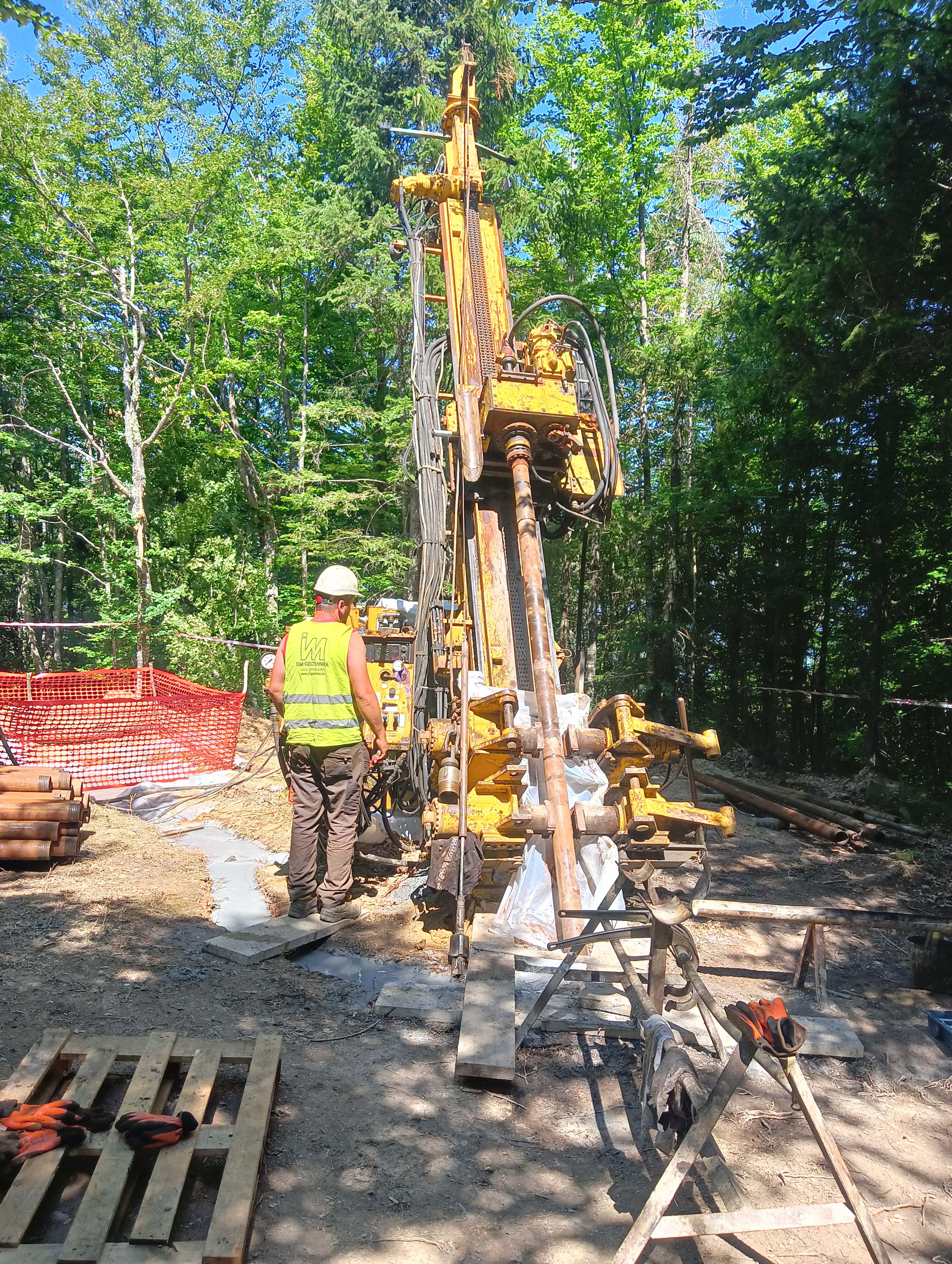 Worker in a hi-vis vest and hard hat operating a yellow diamond drill rig in sunlit forest