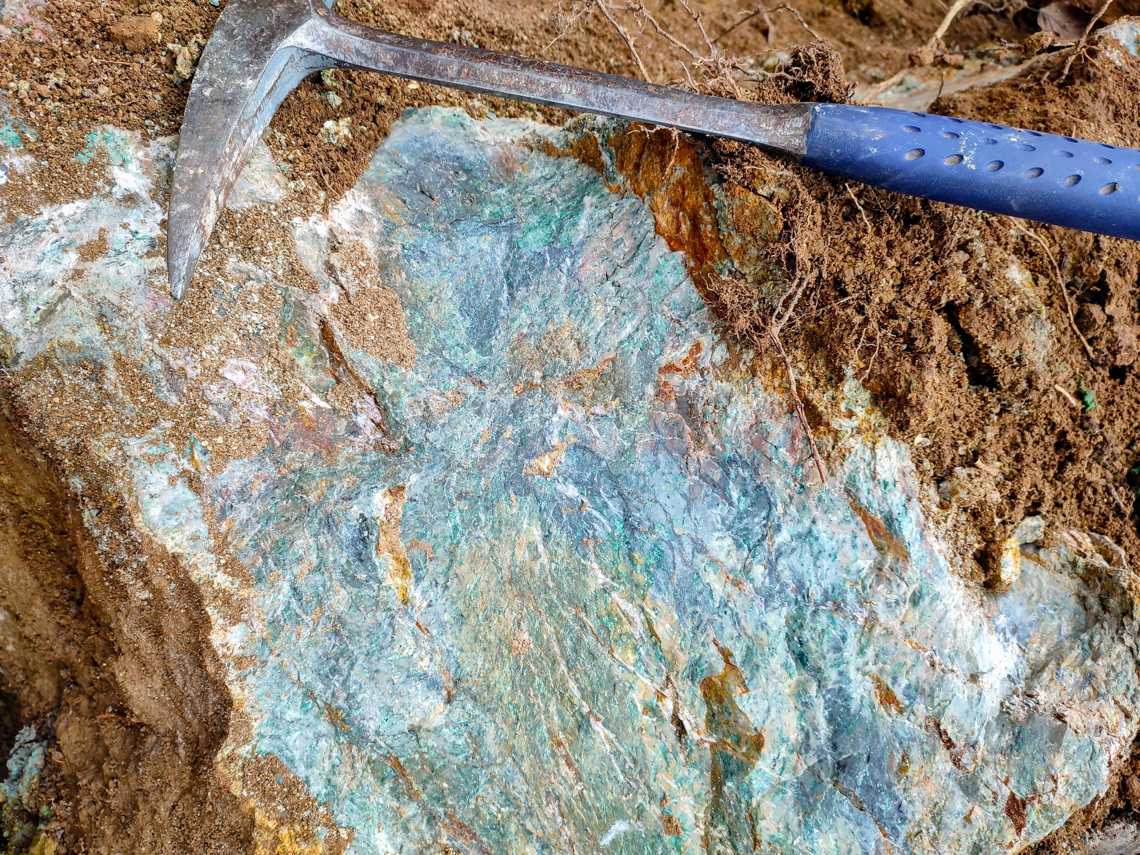 Vivid turquoise-green fuchsite alteration in ultramafic rock exposed in soil, shown with a rock hammer for scale